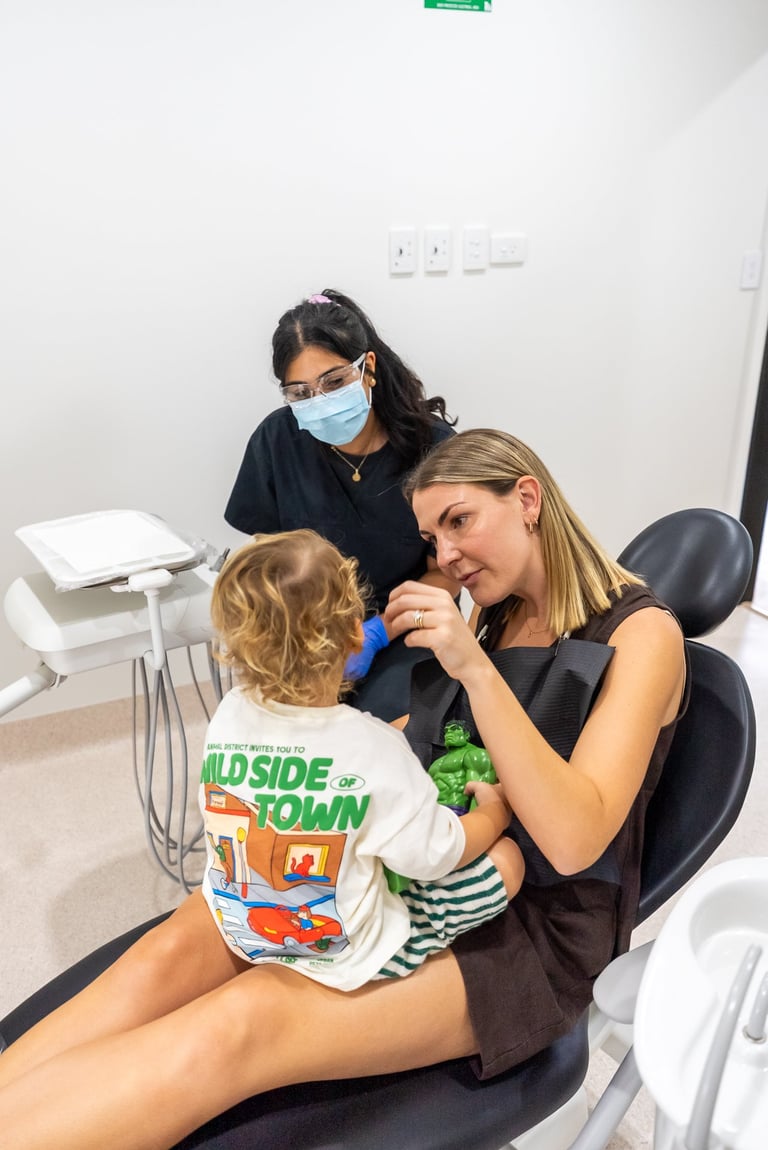 Pediatric dentist in mask examines young childs teeth in dental office with assistant observing