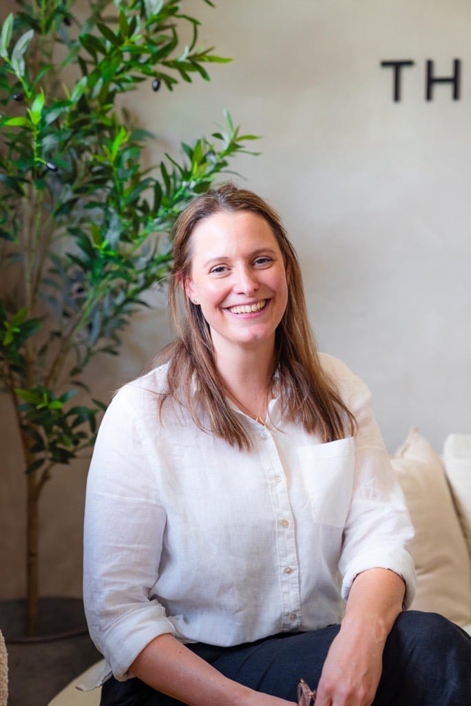 Woman in white button-up shirt smiling at camera, seated in front of green potted plant and beige wall