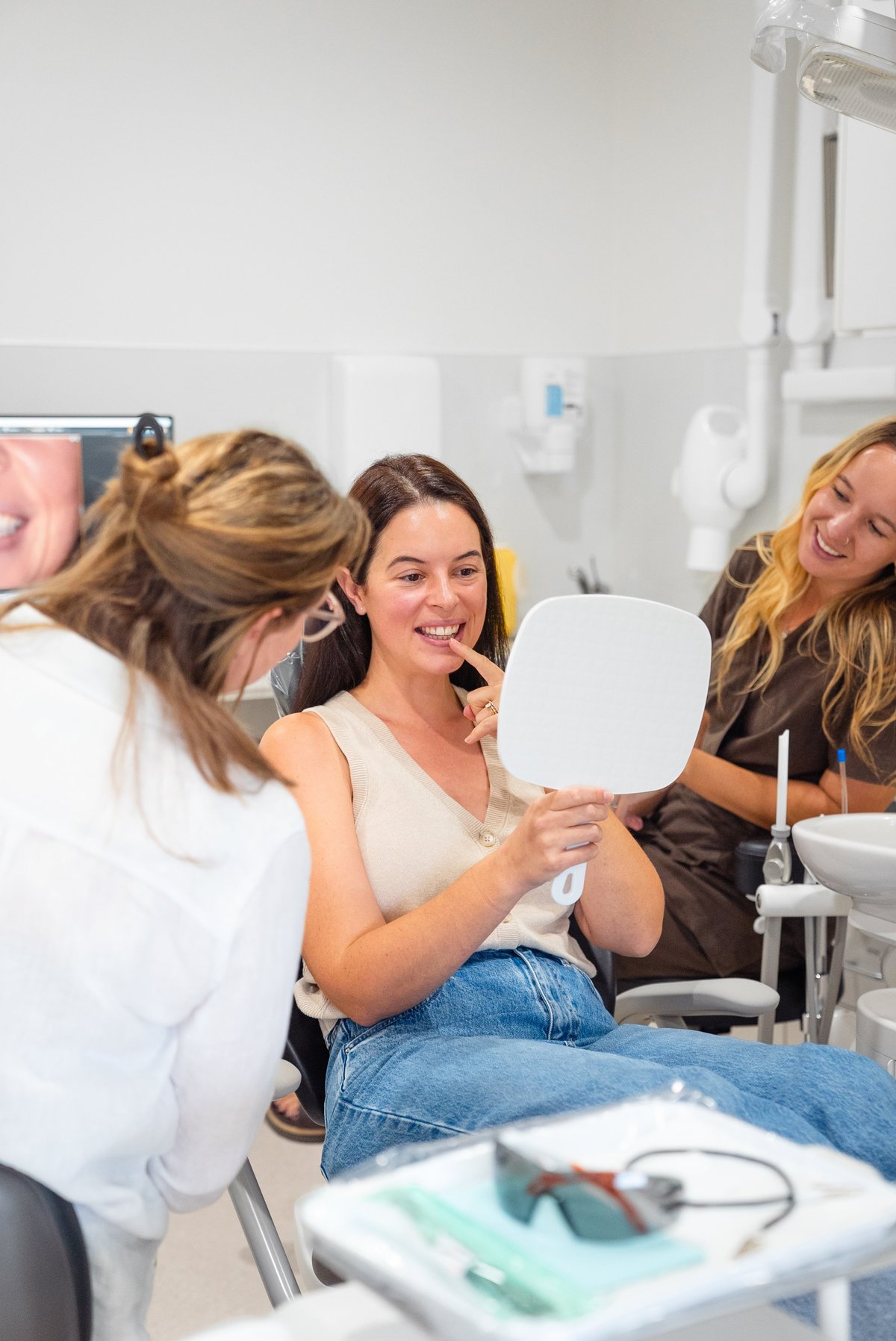 Woman examining her teeth in a mirror at a dental clinic with dentist and staff present