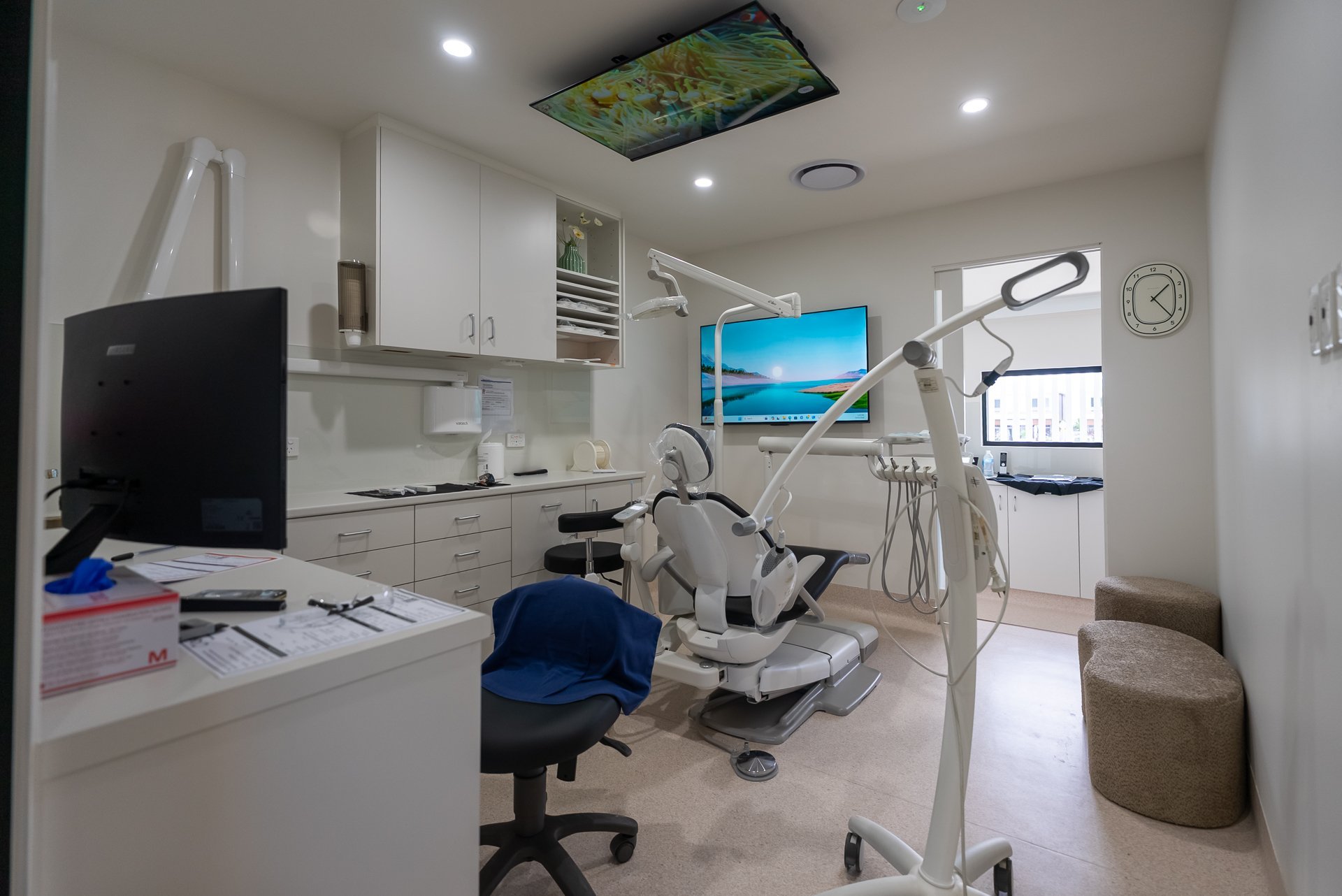 Modern dental office interior with white cabinetry, dental chair, ceiling-mounted light, monitor, and blue rolling stool