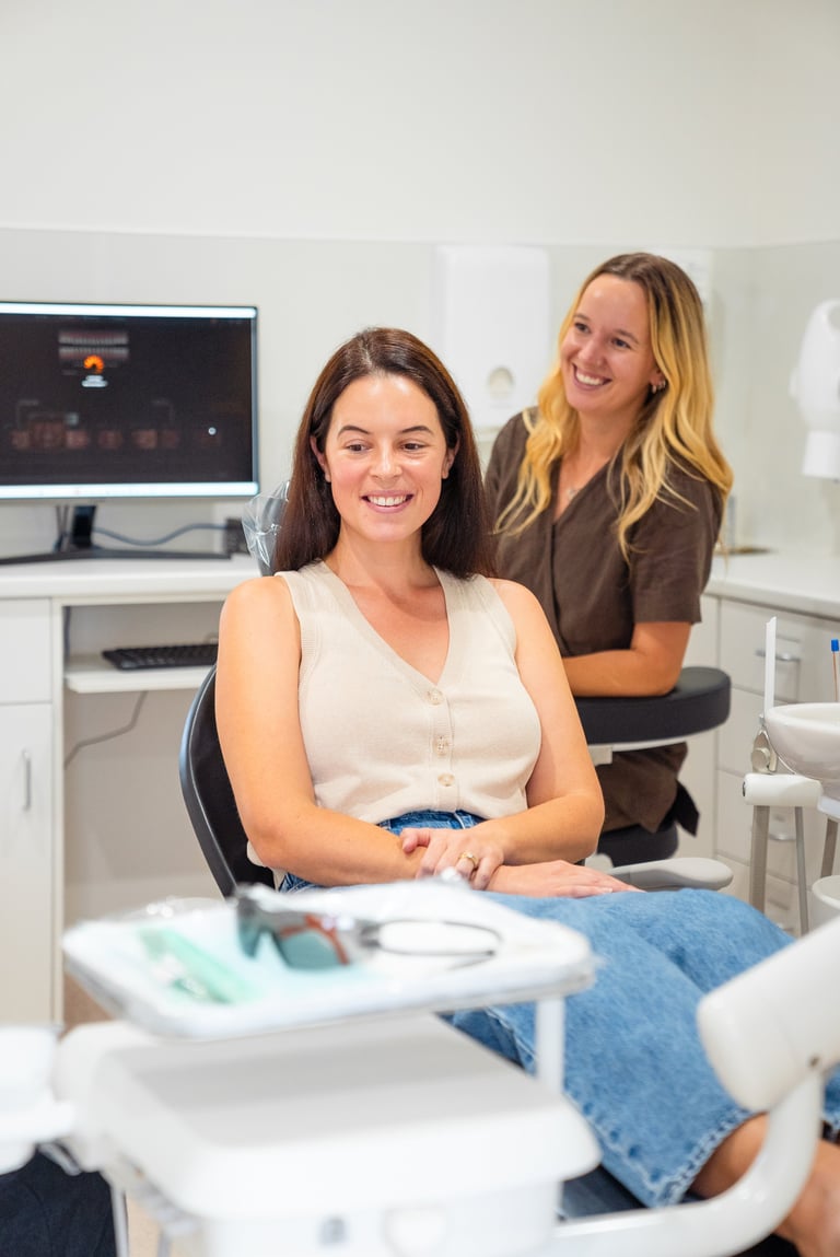Two smiling women in a modern dental office with computer monitor and equipment visible in background