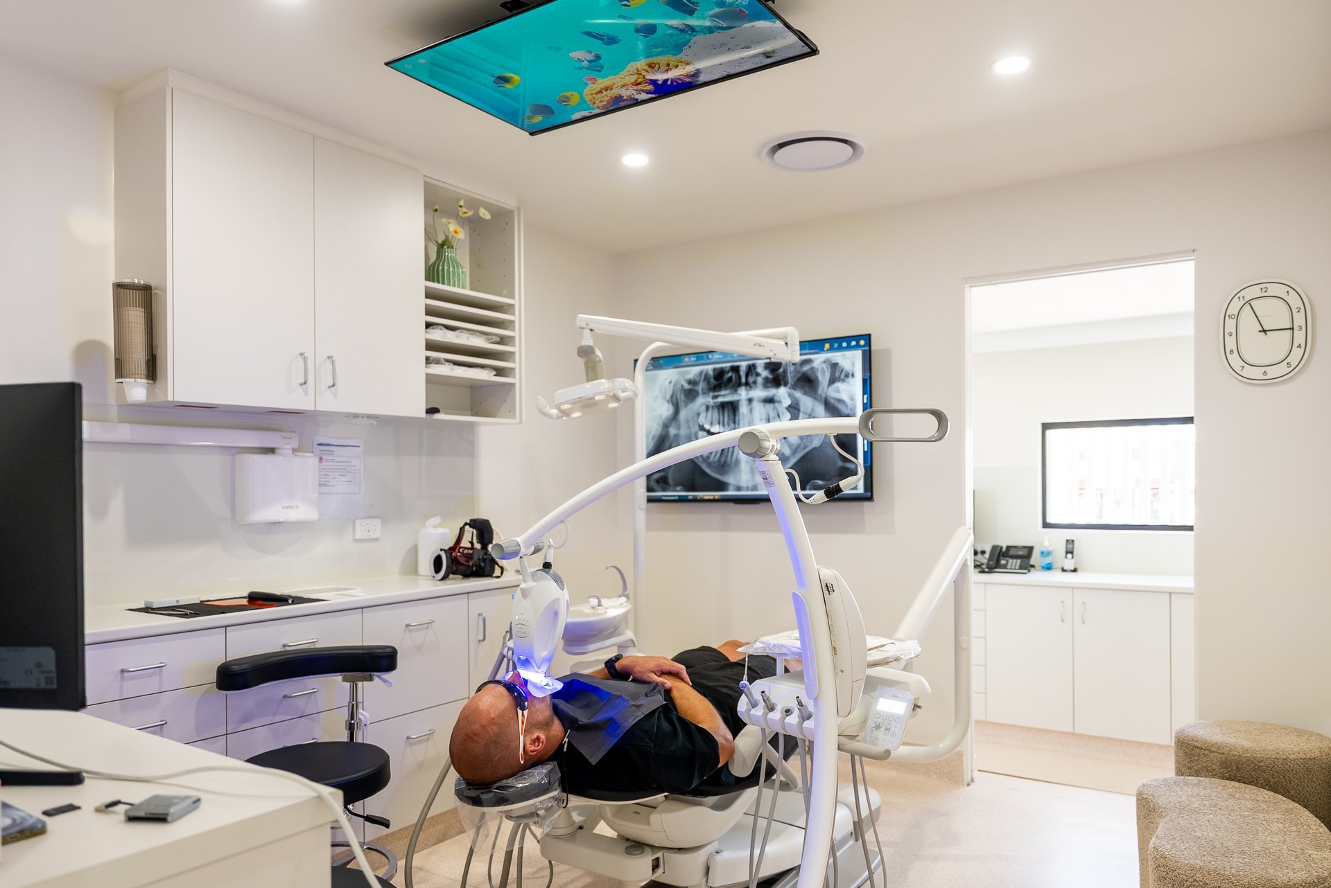 Modern dental office with patient in chair, overhead surgical lights, monitor display, and white cabinetry with equipped workstations