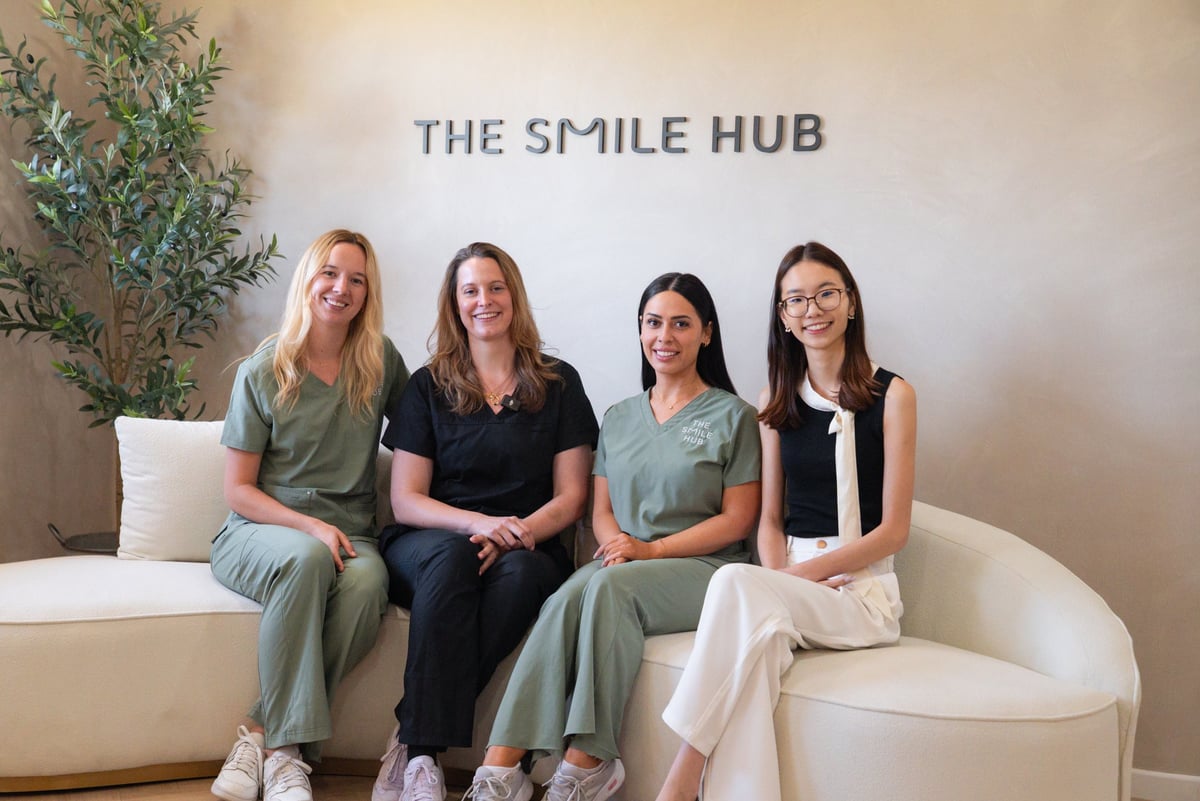 Four women sitting together on a white curved bench in front of a wall displaying THE SMILE HUB text with a potted plant visible on the left