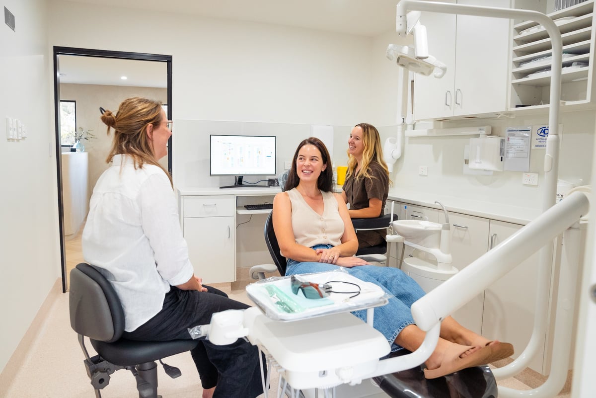 Female dentist consulting with two female patients in a modern dental examination room with white cabinetry and dental equipment.
