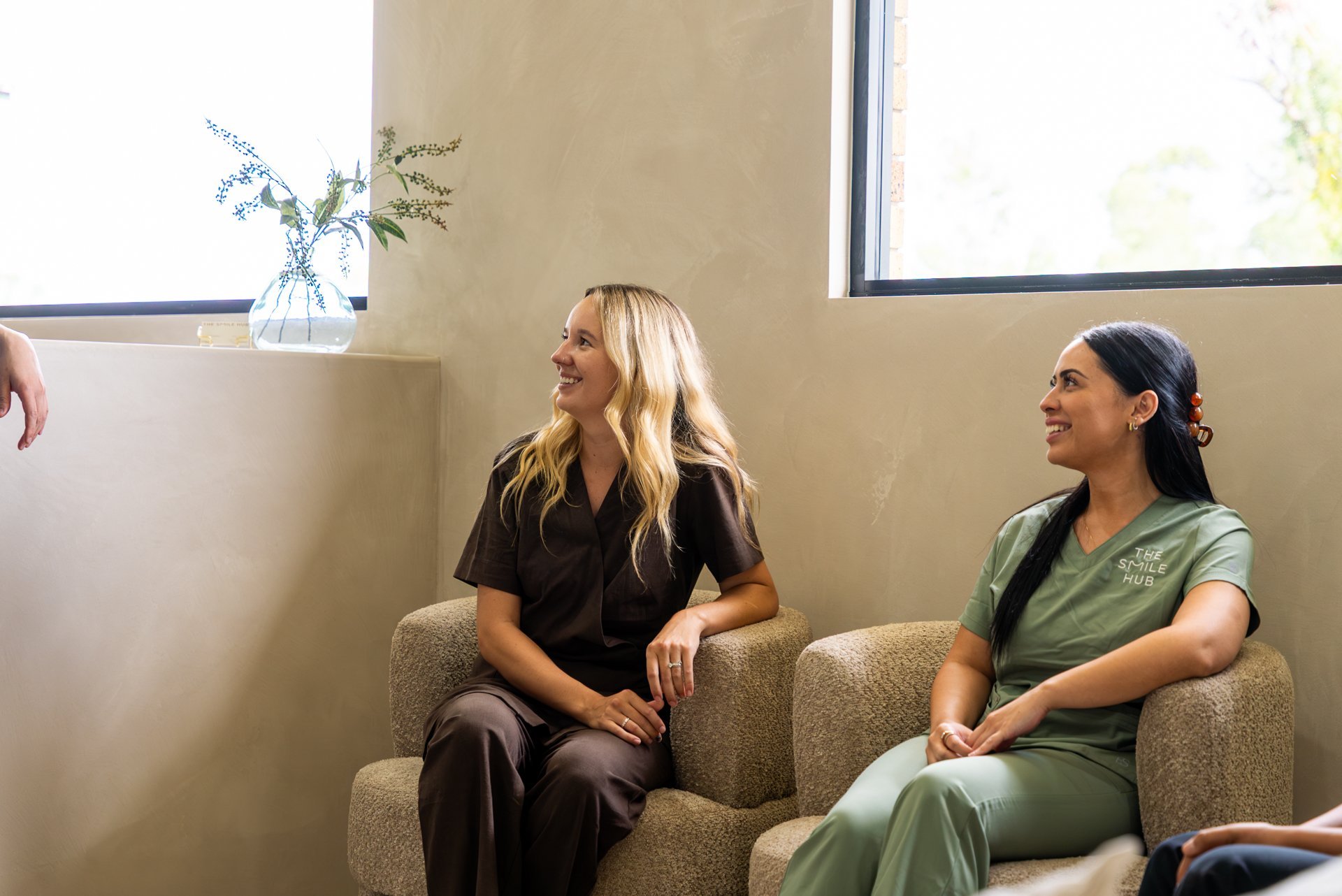 Two women sitting on a beige couch in conversation, with a vase of flowers on a shelf behind them