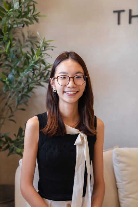 Young woman wearing glasses and black sleeveless top smiling at camera, indoor setting with potted plant behind her